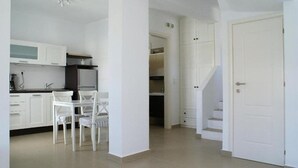 Una cocina moderna con armarios blancos, una zona de comedor con sillas blancas y una escalera que sube a un segundo piso.