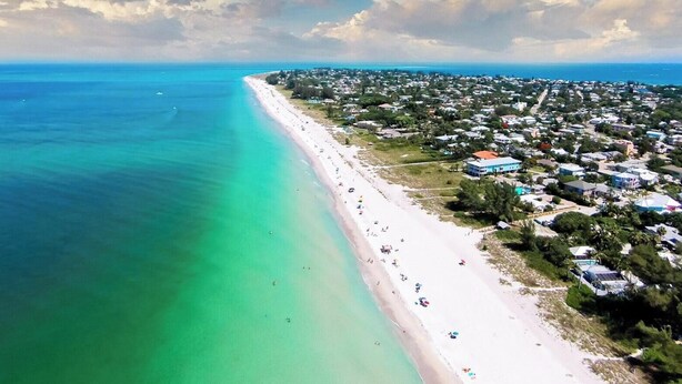 Vista aérea de una zona costera con una extensa playa de arena, edificios residenciales cercanos y aguas azules cristalinas.