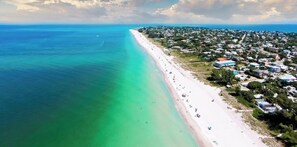 An aerial view of a coastal area with a long sandy beach, adjacent residential buildings, and clear blue waters.
