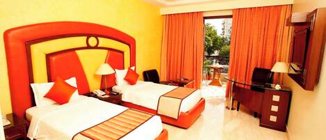 A hotel room with two beds, a desk, a chair, a TV, and a balcony with orange curtains.
