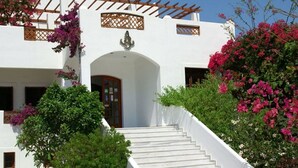 A white building with a staircase leading to an entrance, surrounded by greenery and flowering plants. - Amopi Bay Hotel (Karpathos)