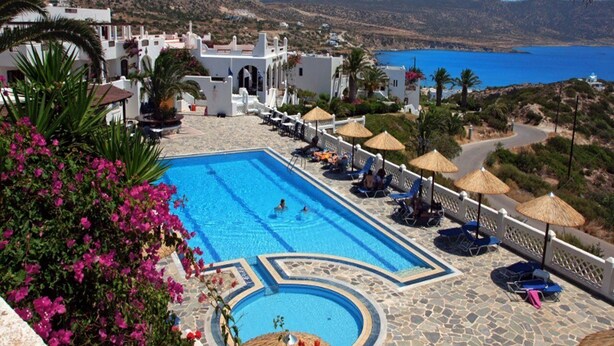 A pool area with a view of the sea, surrounded by white buildings and palm trees. - Amopi Bay Hotel (Karpathos)