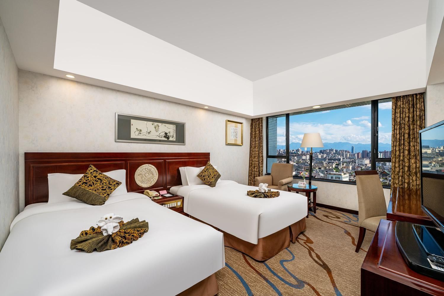 Ein Hotelzimmer mit zwei Betten, einem Schreibtisch, einem Sessel, einem Fernseher und Blick auf die Stadt.