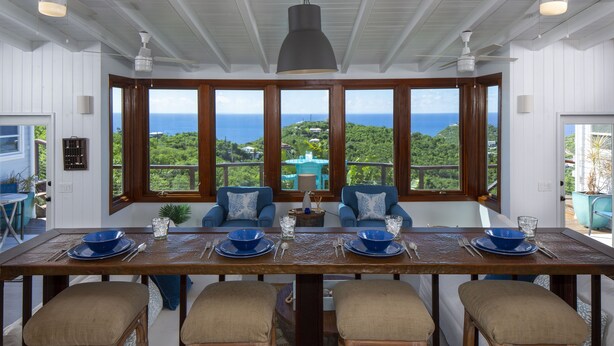 A dining area with a view of the ocean, a large table with blue and beige chairs, and a ceiling fan. - Stunning Views, Peaceful Location, Large Pool & Minutes from Cruz Bay (cruz bay)
