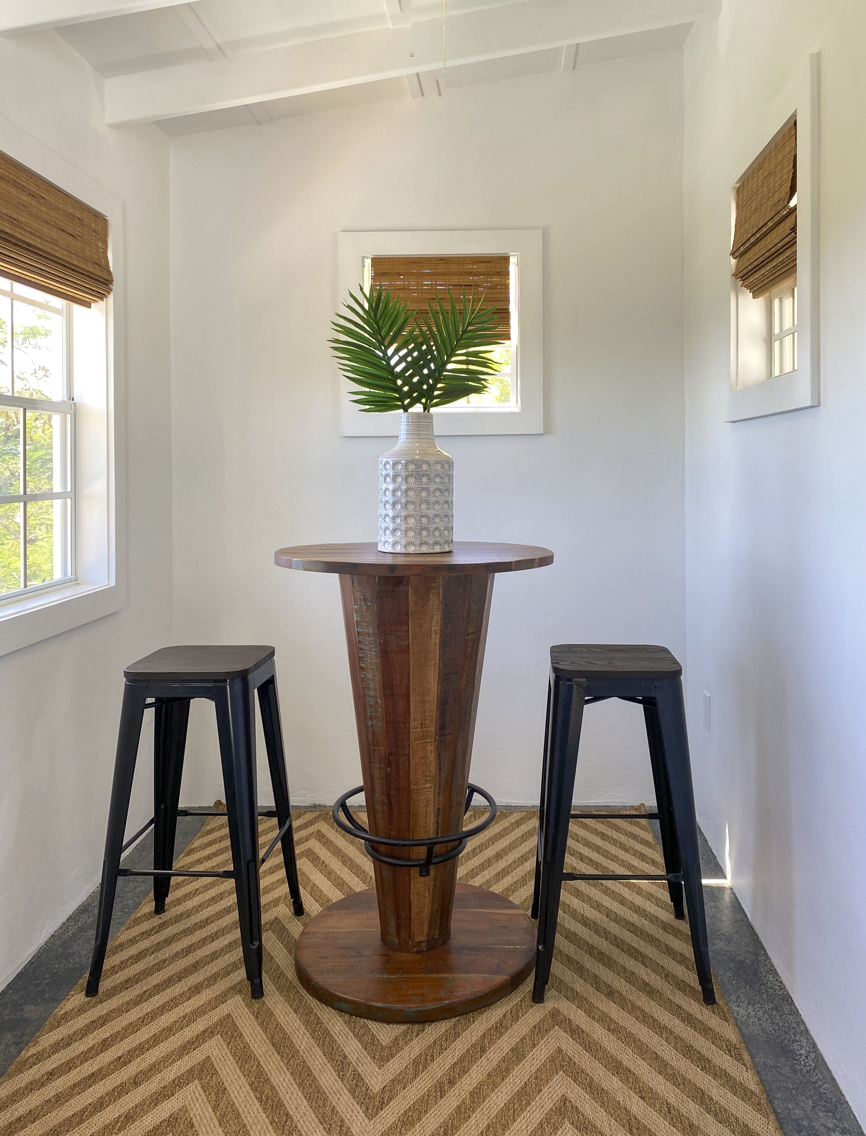 A corner room with a wooden table, two black stools, a potted plant, and a window with blinds.