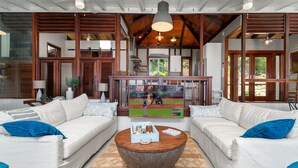 A spacious living room with two white sofas, a wooden coffee table, and a large flat-screen TV displaying a baseball game. - Stunning Views, Peaceful Location, Large Pool & Minutes from Cruz Bay (cruz bay)