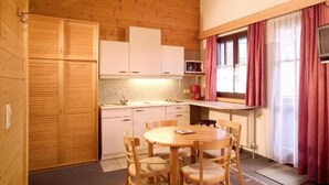 A compact kitchen with wooden cabinets, a dining table with chairs, and a window with curtains. - Appartmenthaus Raudaschl (Laengenfeld)