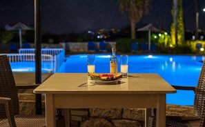 A table with a bottle and glasses, a plate with food, and a pool in the background.
