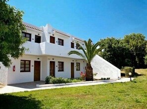 A white two-story building with a flat roof, multiple windows, and a small garden area with a palm tree.