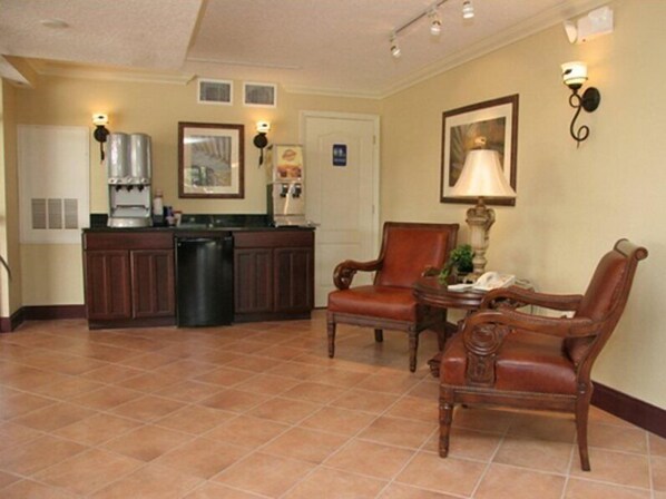 A kitchen with a coffee maker, refrigerator, and a trash bin. There are two wooden chairs and a table with a lamp.