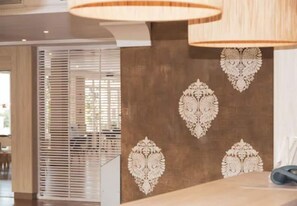 A room with a wooden floor, a wall with decorative patterns, a window with blinds, and a table with a lamp.