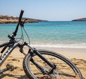Uma bicicleta estacionada numa praia de areia, com vista para o mar e colinas ao longe.