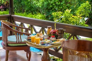 A wooden chair with a cushion, a table with breakfast items, and a view of greenery. - Villa Chempaka (Ubud)