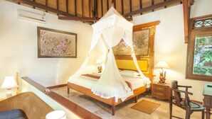 A bedroom with a four-poster bed, a wooden headboard, a bedside table with a lamp, a chair, and a view of greenery through a window. - Villa Chempaka (Ubud)
