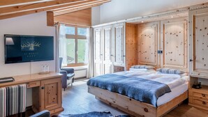 Ein Schlafzimmer mit einem hölzernen Bett, einem Schreibtisch und einem Stuhl. Es gibt ein Fenster mit Vorhängen und einen an der Wand befestigten Fernseher.