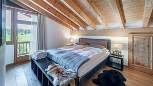 Ein Schlafzimmer mit Holzbalkendecke, einem großen Bett, einem Nachttisch, einem Kleiderschrank und Blick auf eine grüne Landschaft durch ein Fenster.