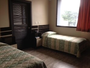 Una habitación con dos camas, una mesita de noche, un ventanal con cortinas y una puerta con persianas.