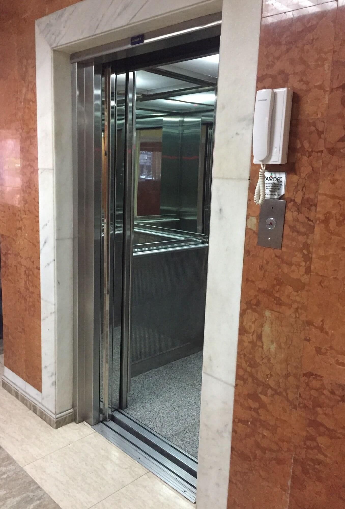 An elevator with a glass door, a wall-mounted intercom, and a tiled floor.