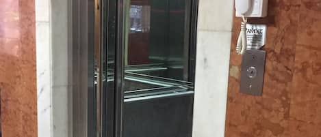 An elevator with a glass door, a wall-mounted intercom, and a tiled floor.