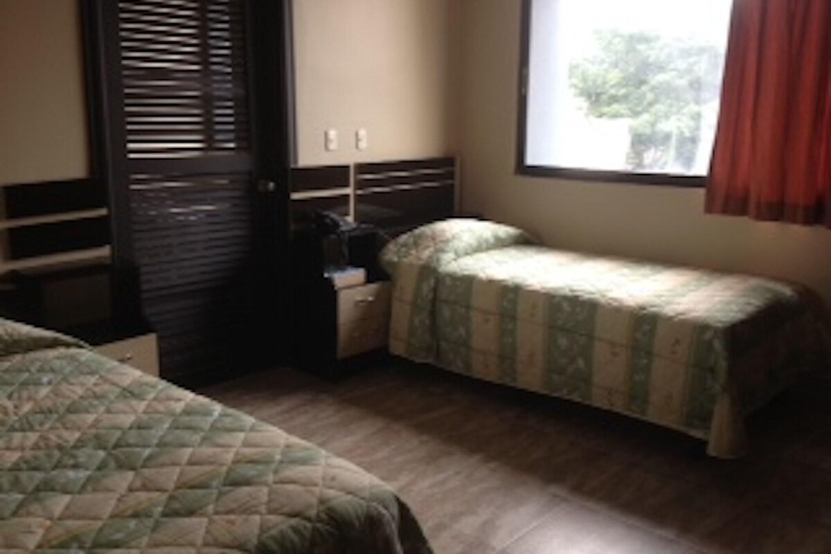 A room with two beds, a nightstand, a window with curtains, and a door with blinds.