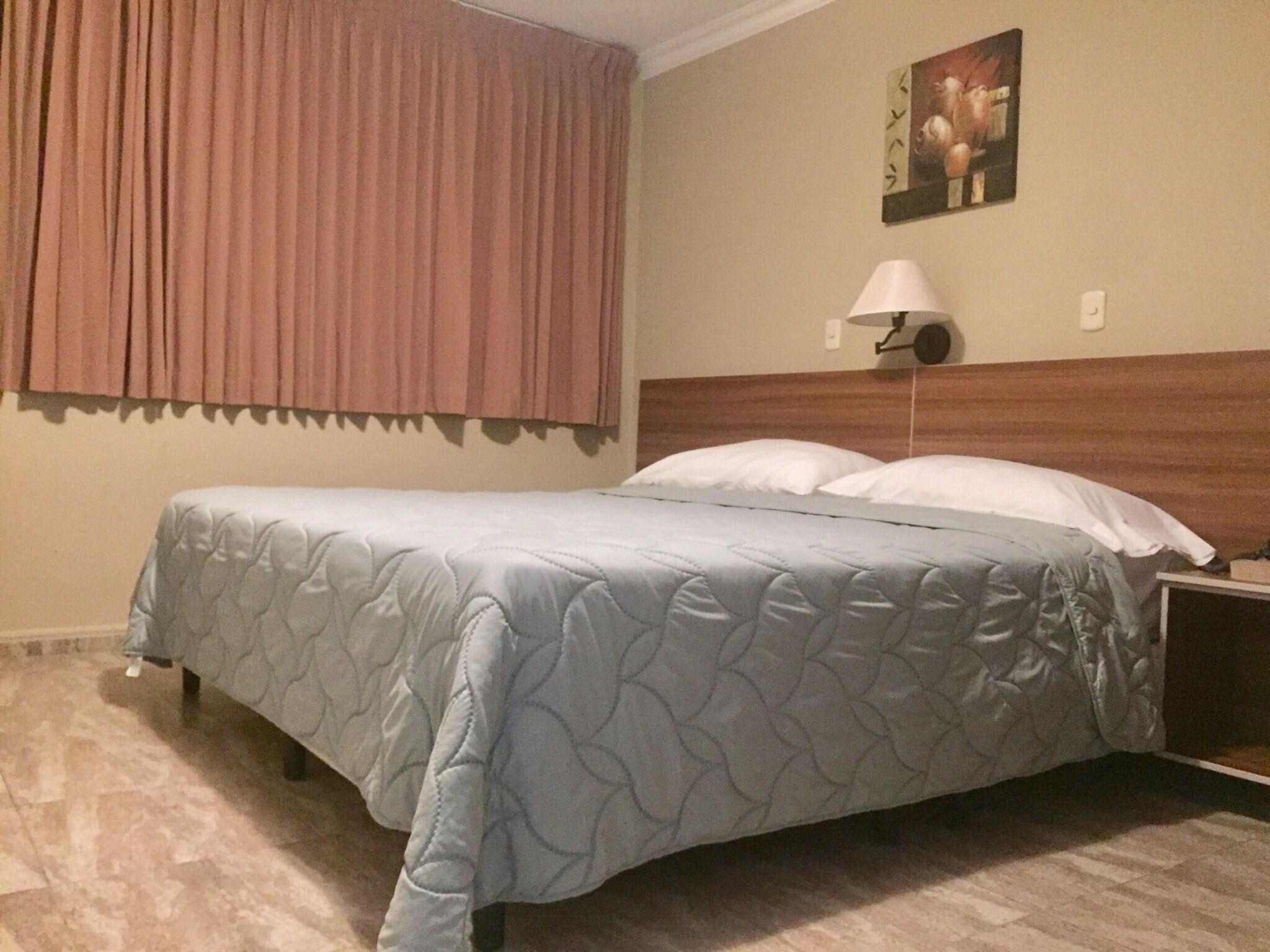 A hotel room with a bed, a nightstand, a lamp, a painting on the wall, and a window with curtains.