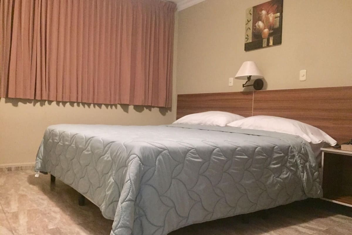 Ein Hotelzimmer mit Bett, Nachttisch, Lampe, einem Gemälde an der Wand und einem Fenster mit Vorhängen.