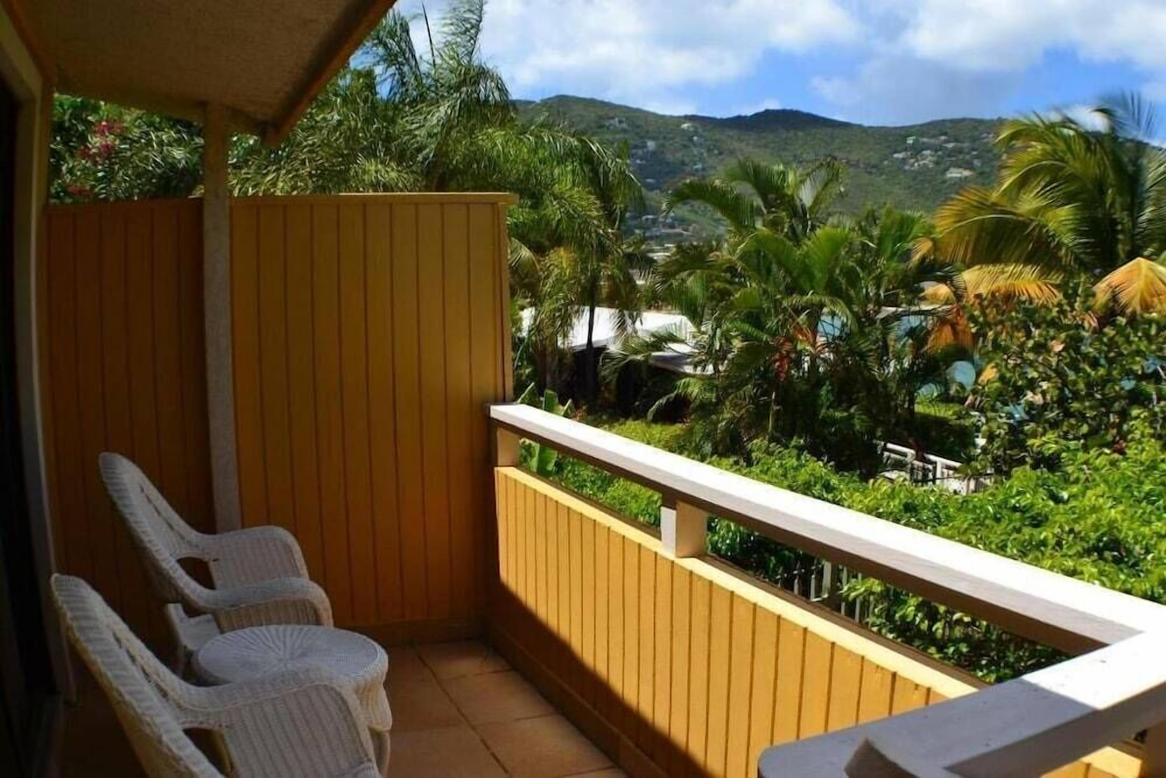 A balcony with wicker chairs, a wooden railing, and a view of lush greenery and distant hills.