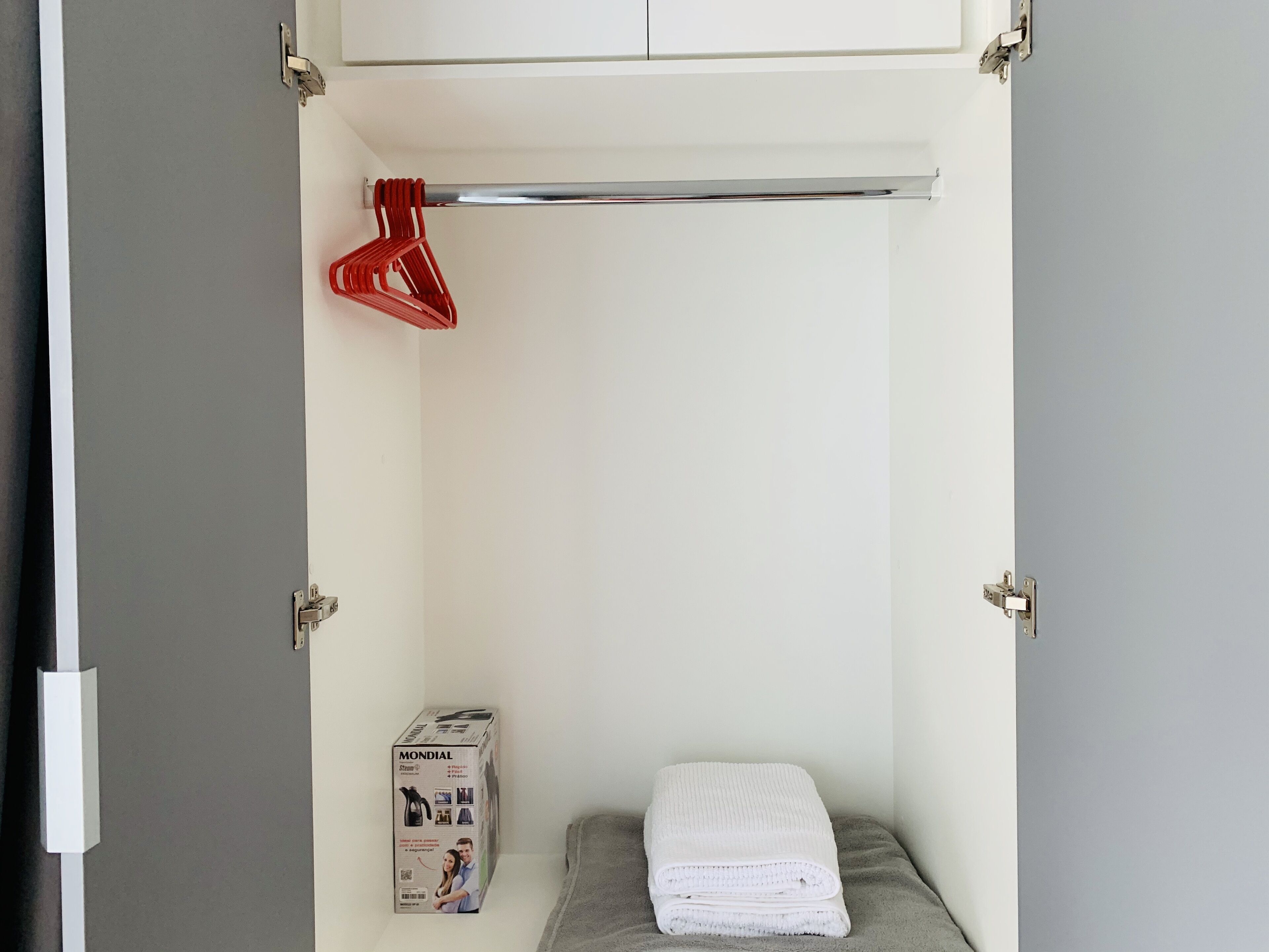 Een kleine, netjes georganiseerde kast met een witte handdoek gevouwen op een grijze plank, een rode kleerhanger, en een doos van een product.