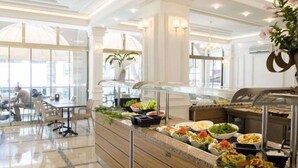 A buffet area with various food options, a seating area with tables and chairs, and a large window providing natural light. - Ozcan Hotel (Marmaris)