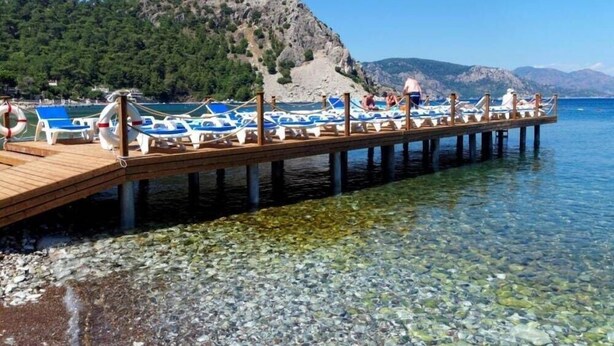 A wooden pier with blue lounge chairs and boats, leading to a clear, rocky shoreline with mountains in the background. - Ozcan Hotel (Marmaris)