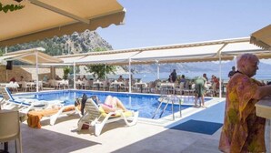 A poolside area with lounge chairs, umbrellas, and tables under a canopy, overlooking a body of water and rocky terrain. - Ozcan Hotel (Marmaris)