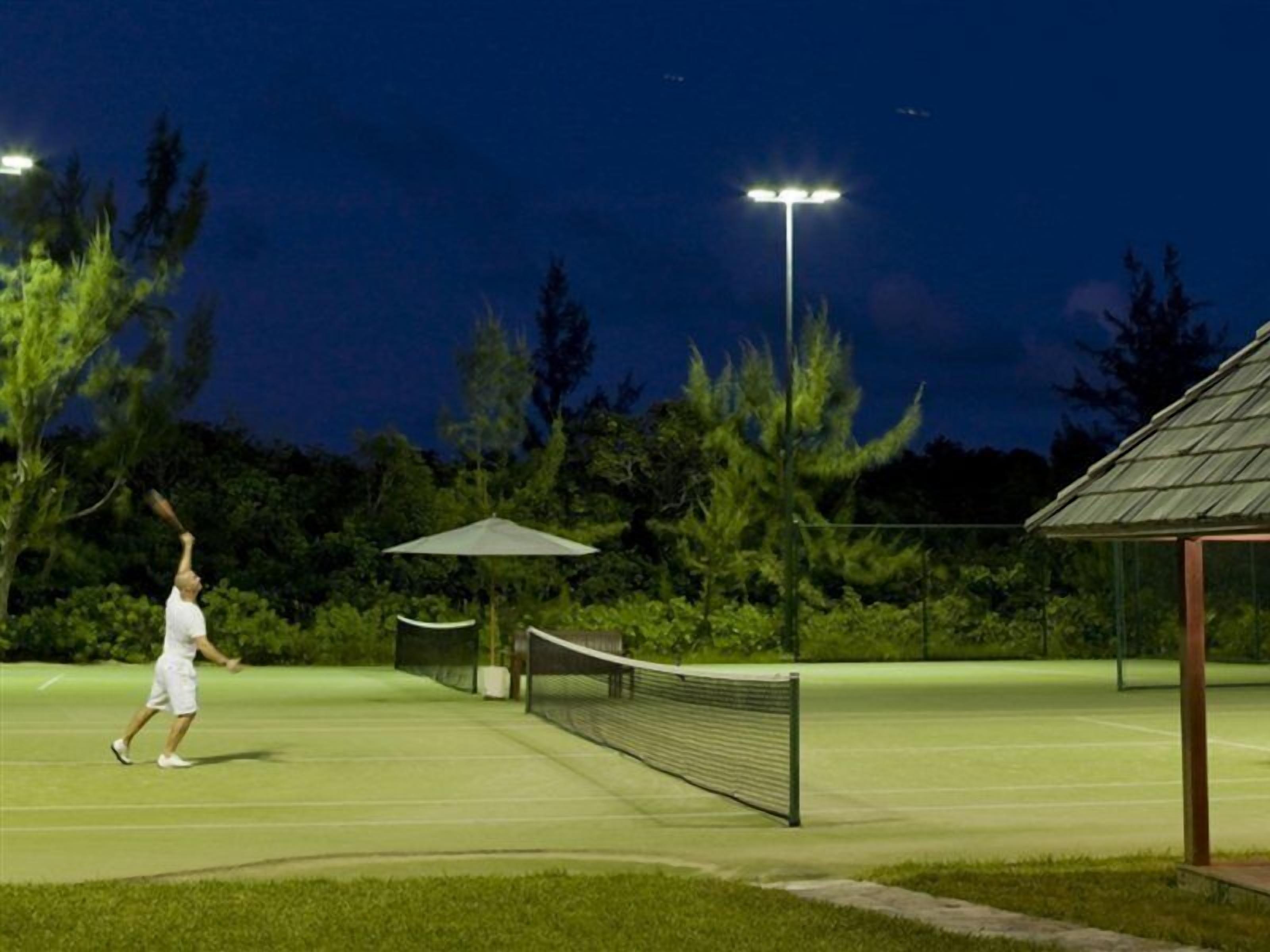 A tennis court at night with a net, a bench, and a gazebo in the background.