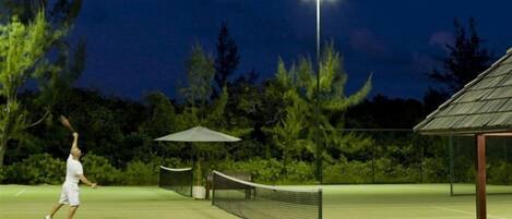 A tennis court at night with a net, a bench, and a gazebo in the background.