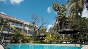 A swimming pool with a clear blue sky, palm trees, and a multi-story building with balconies.