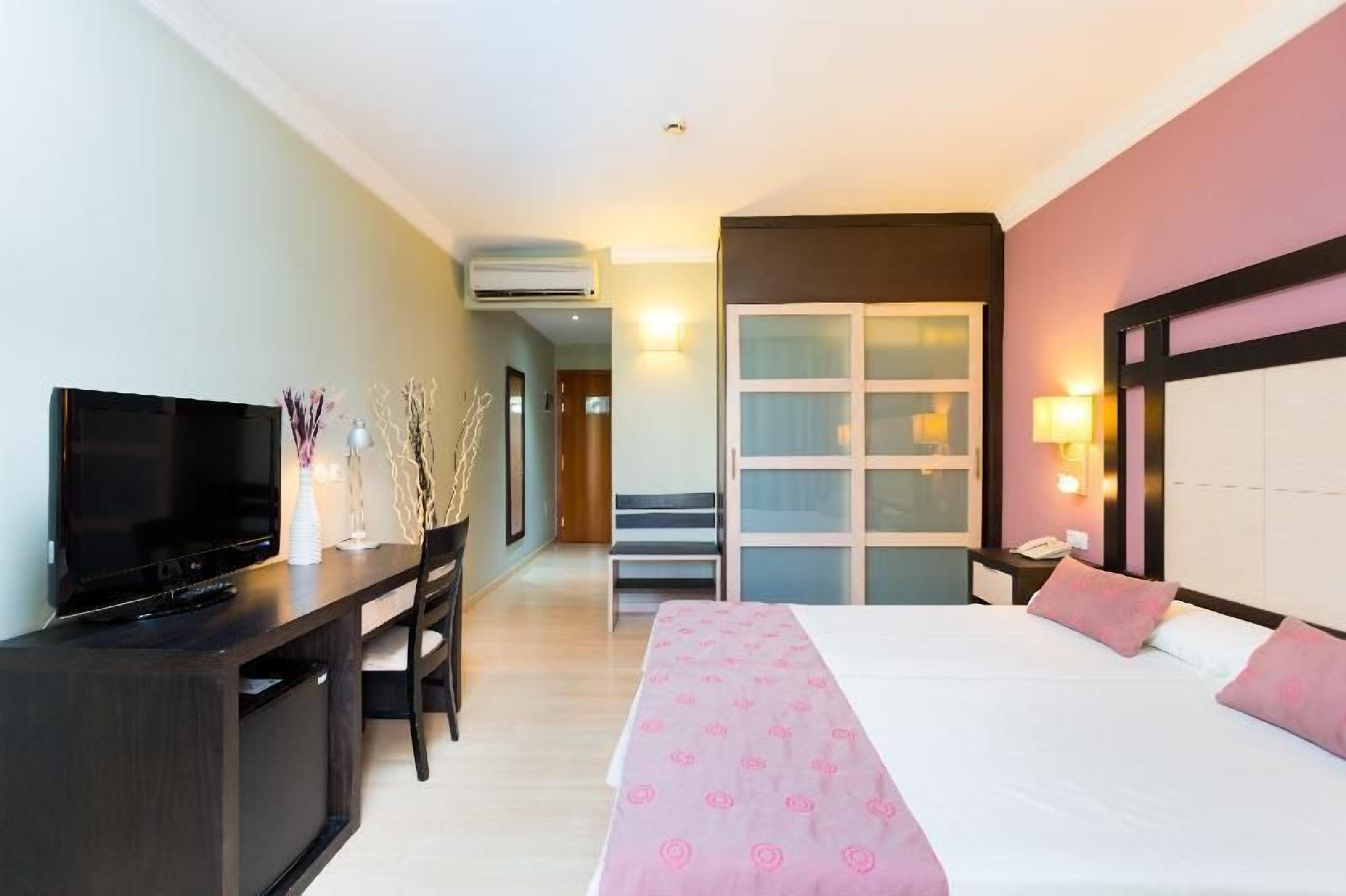 A hotel room with a bed, a desk with a television, a chair, a nightstand, and a wardrobe.