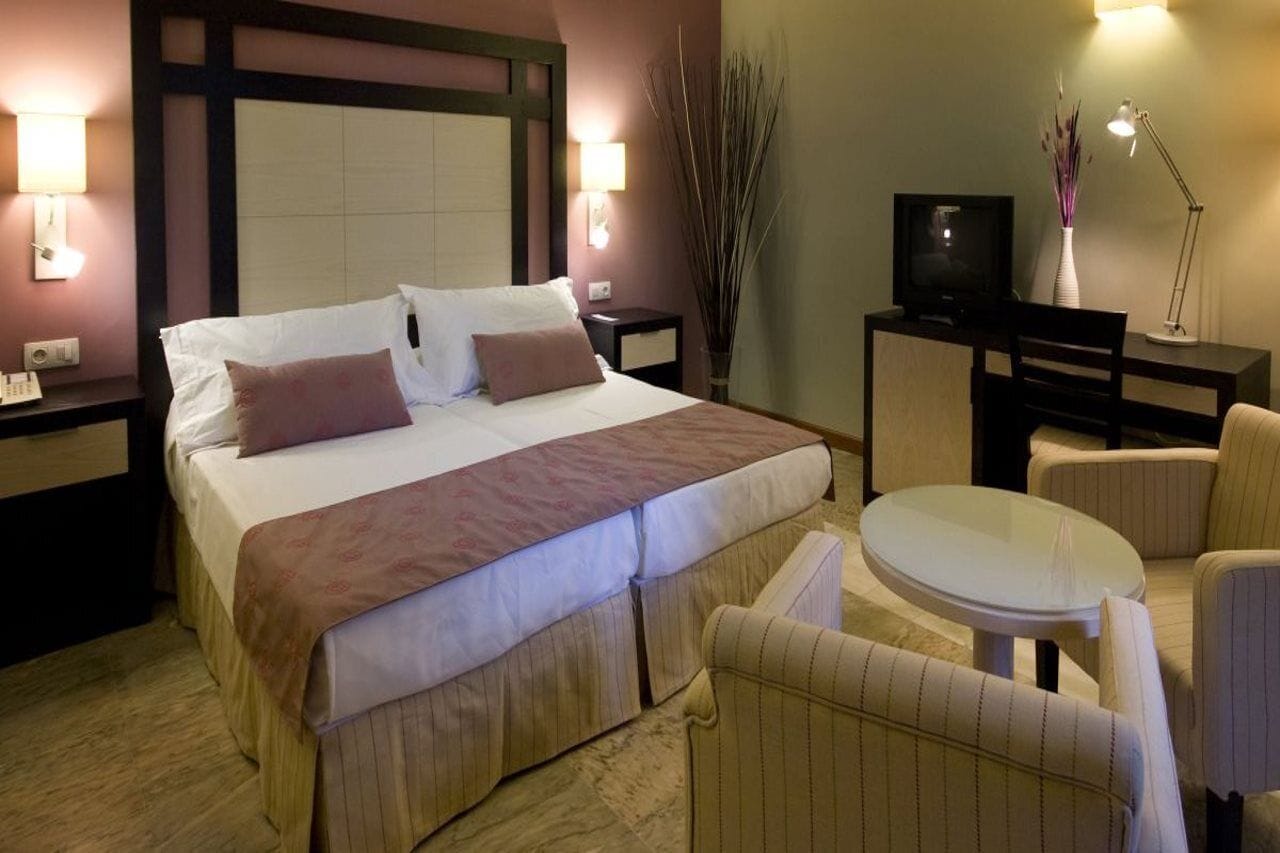 Una habitación de hotel con cama, mesitas de noche, televisión, escritorio y una mesa redonda con sillas.