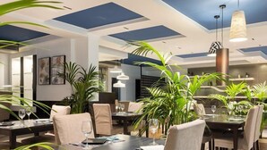 A modern dining area with multiple tables and chairs, large potted plants, and contemporary lighting fixtures.
