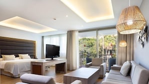 A modern hotel room with a large bed, a desk with a flat-screen TV, a sofa, and a balcony with a view of greenery.