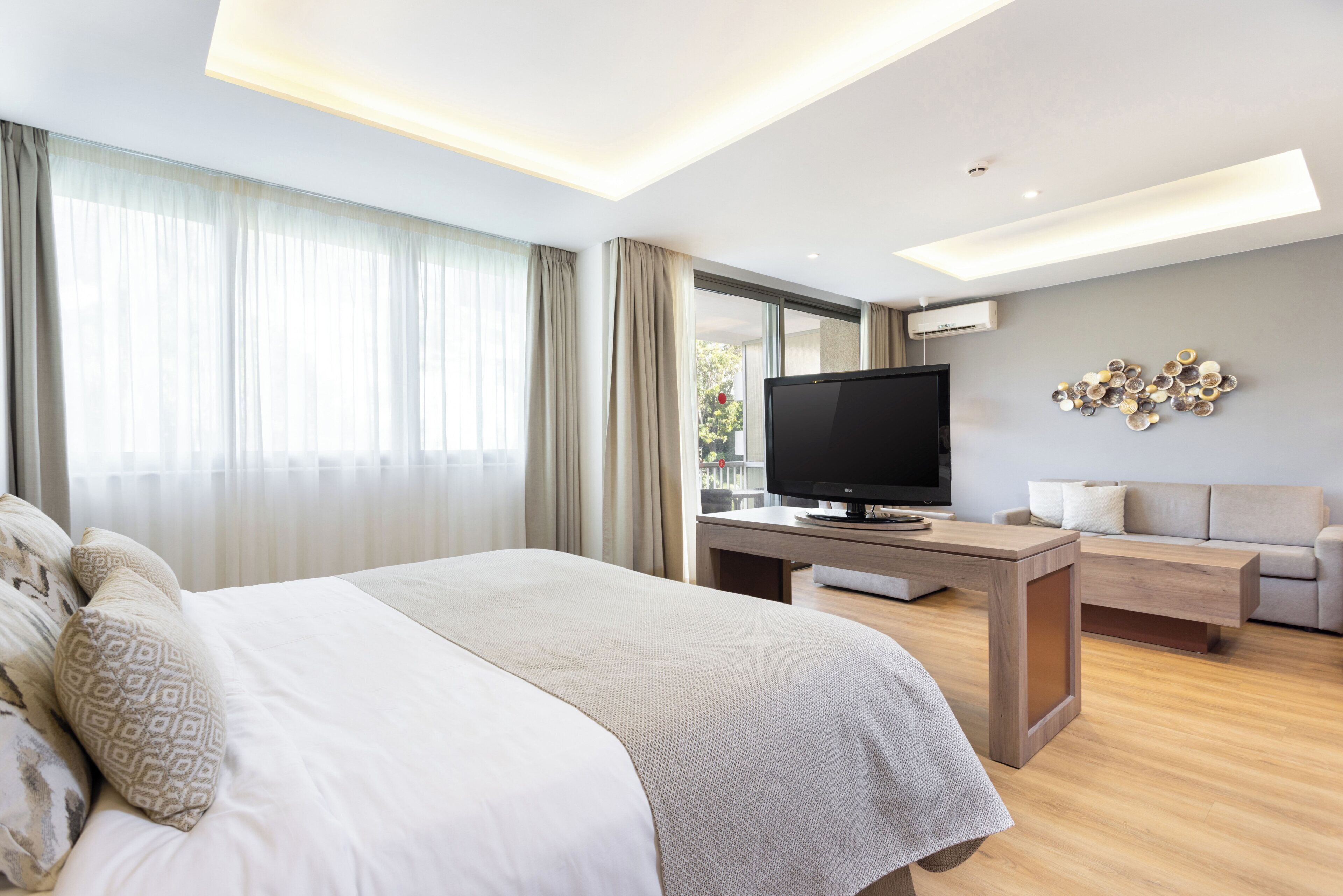 Quarto de hotel moderno com uma cama grande, televisão de tela plana, sofá e um objeto decorativo na parede.