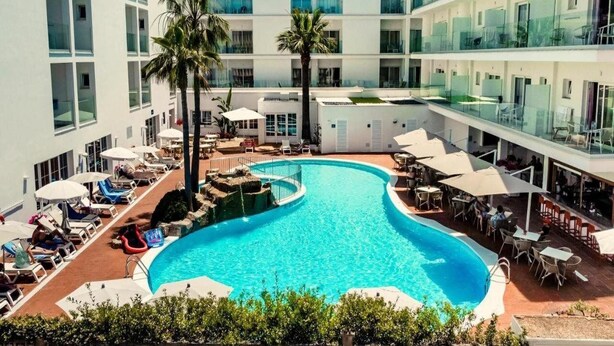 A modern hotel with a central swimming pool surrounded by lounge chairs and umbrellas, multiple floors with balconies, and a clear sky. - Hotel Capricho (Capdepera)