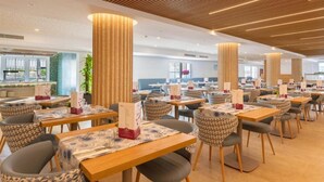 A modern dining area with wooden tables and chairs, large cylindrical columns, and a bright ceiling with linear lighting. - Hotel Capricho (Capdepera)