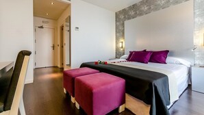 A modern hotel room with a large bed, two pink ottomans, a wooden bench, and a stone accent wall. - YIT Via Sevilla Mairena (Mairena del Aljarafe)