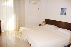 Una cama bien hecha con cabecera de madera, una mesita de noche con lámpara y una ventana con cortinas traslúcidas.