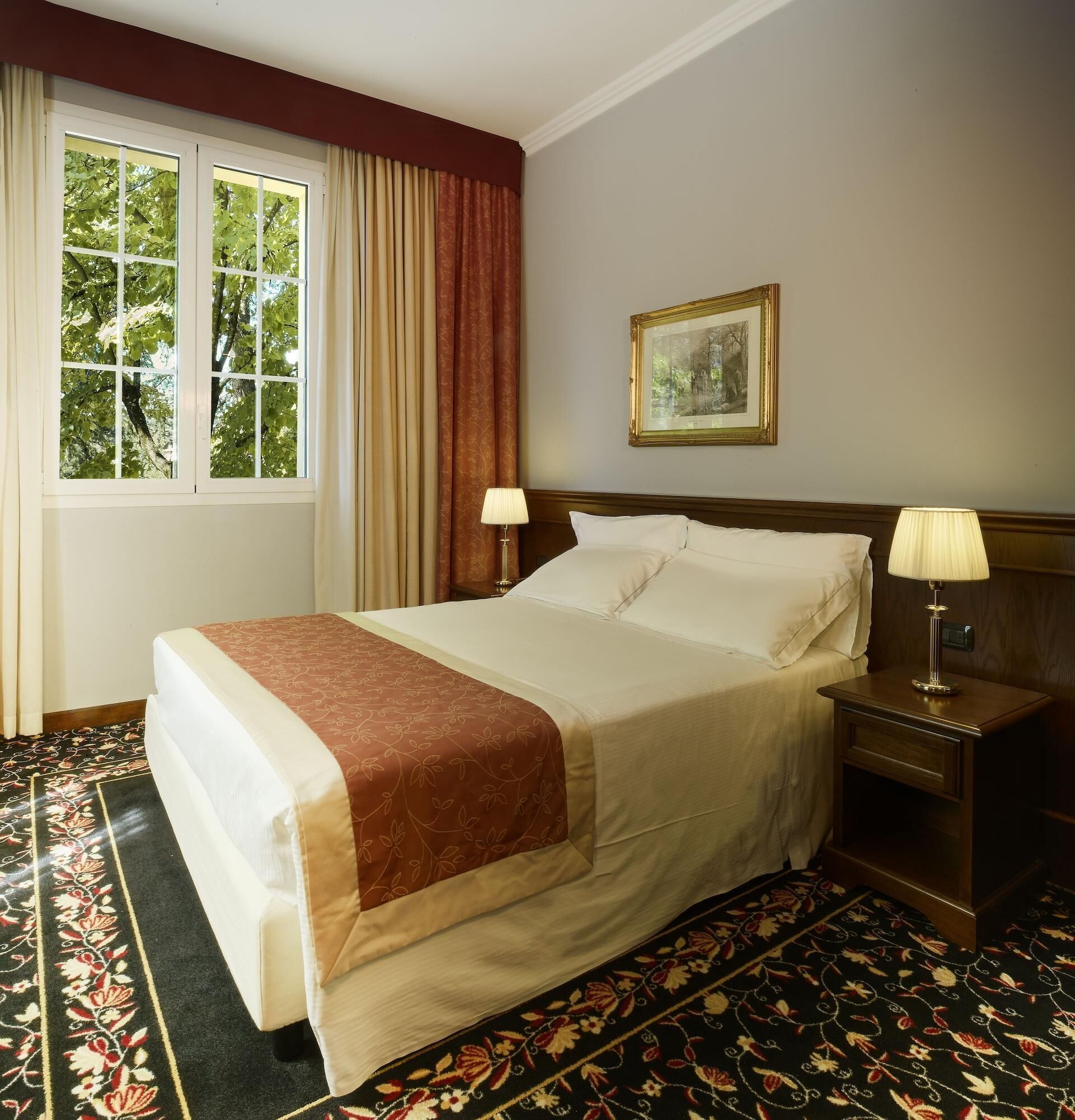 A neatly made bed with a floral bedspread, two bedside lamps, a framed picture on the wall, and a window with curtains.