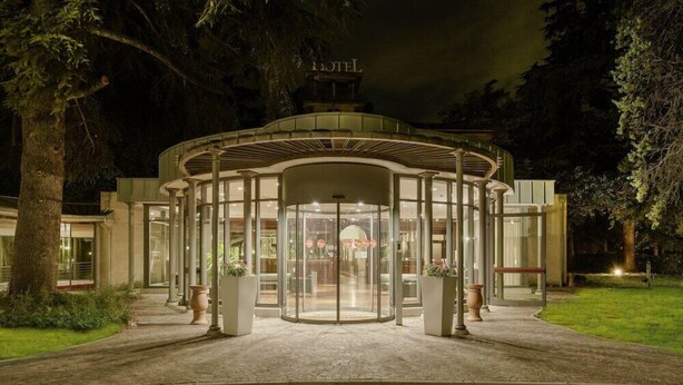 A hotel entrance with a glass revolving door, surrounded by trees and a well-lit pathway. - Meditur Hotel Bologna (San Lazzaro di Savena)