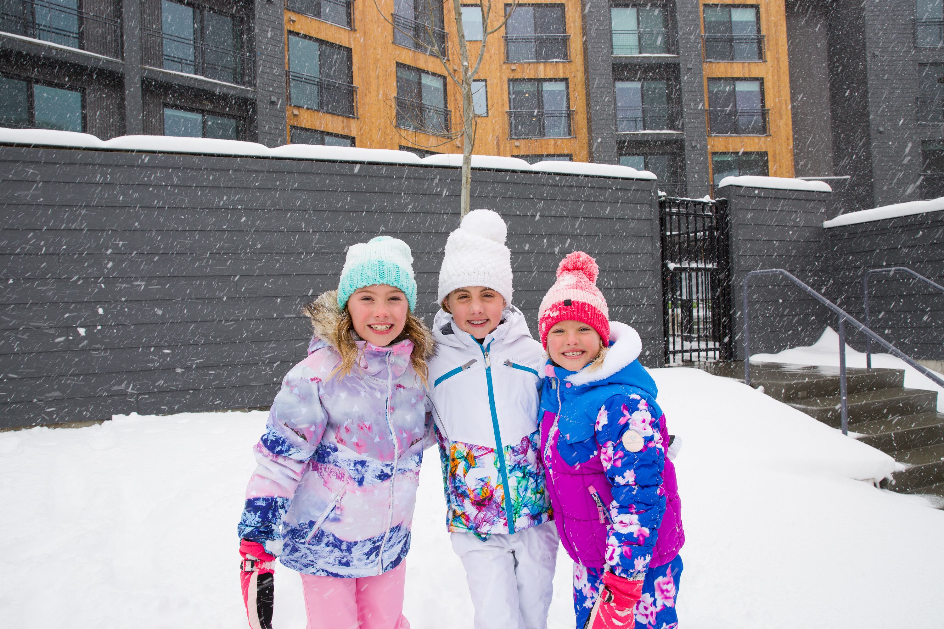 Tre bambini in piedi in una zona innevata, con un edificio sullo sfondo.