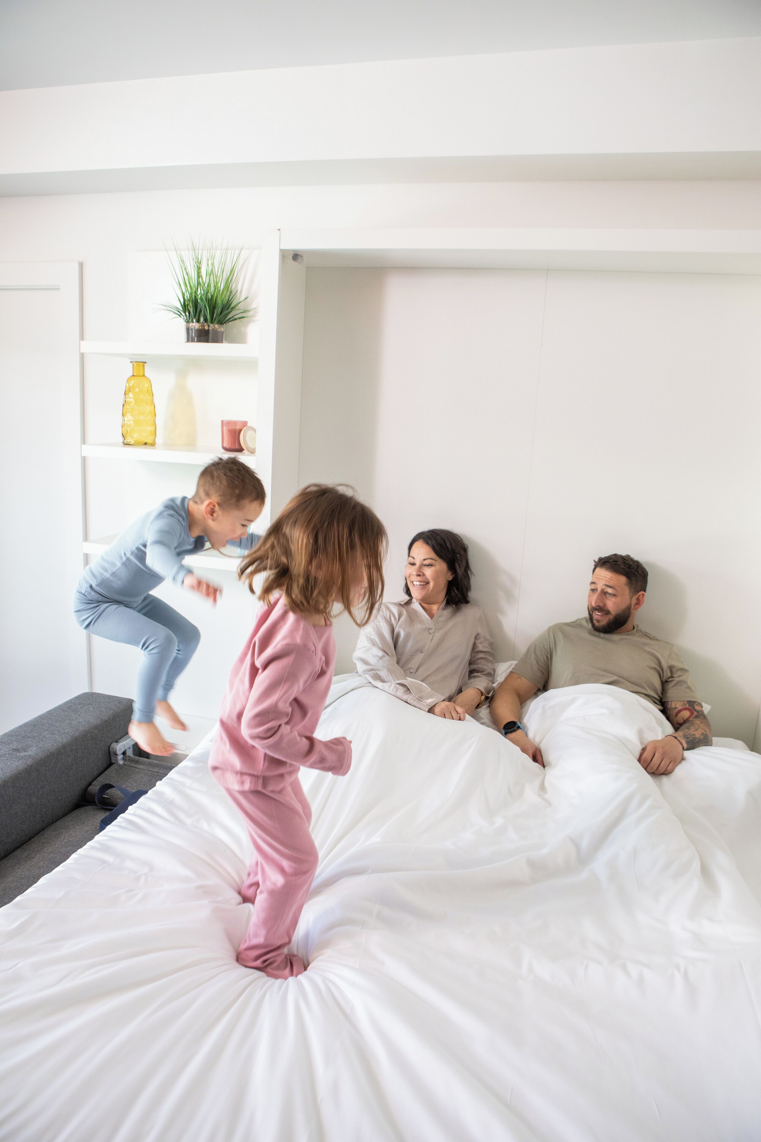 A family in a bedroom with a bed, a shelf with items, and a plant.