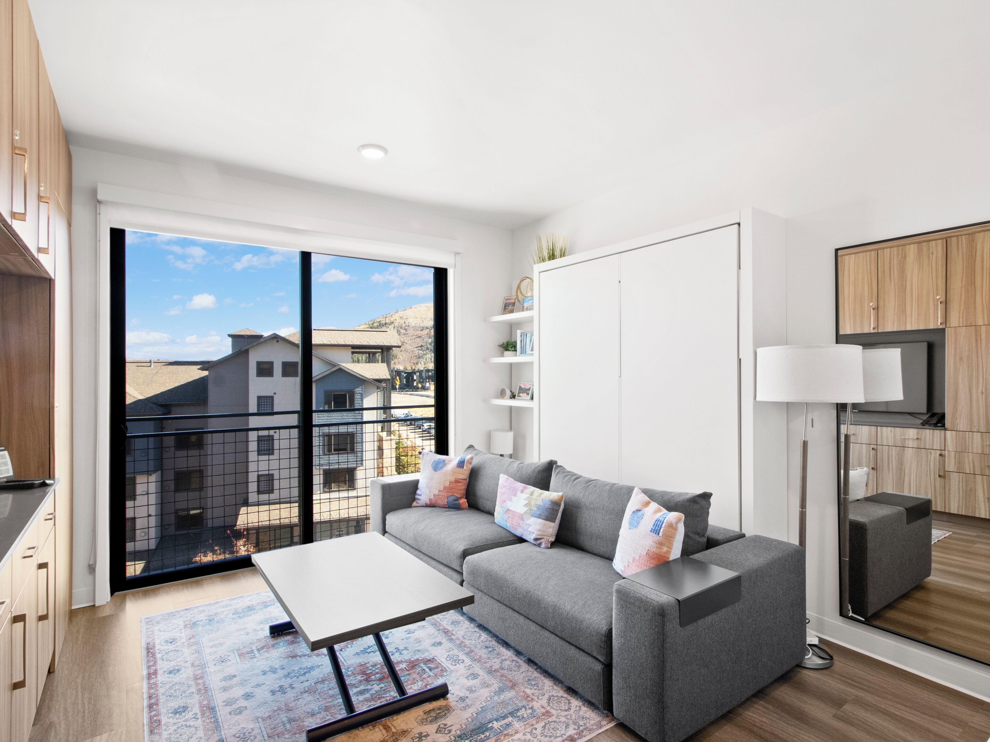 A modern living room with a grey sofa, a small table, and a view of a residential area through large windows.