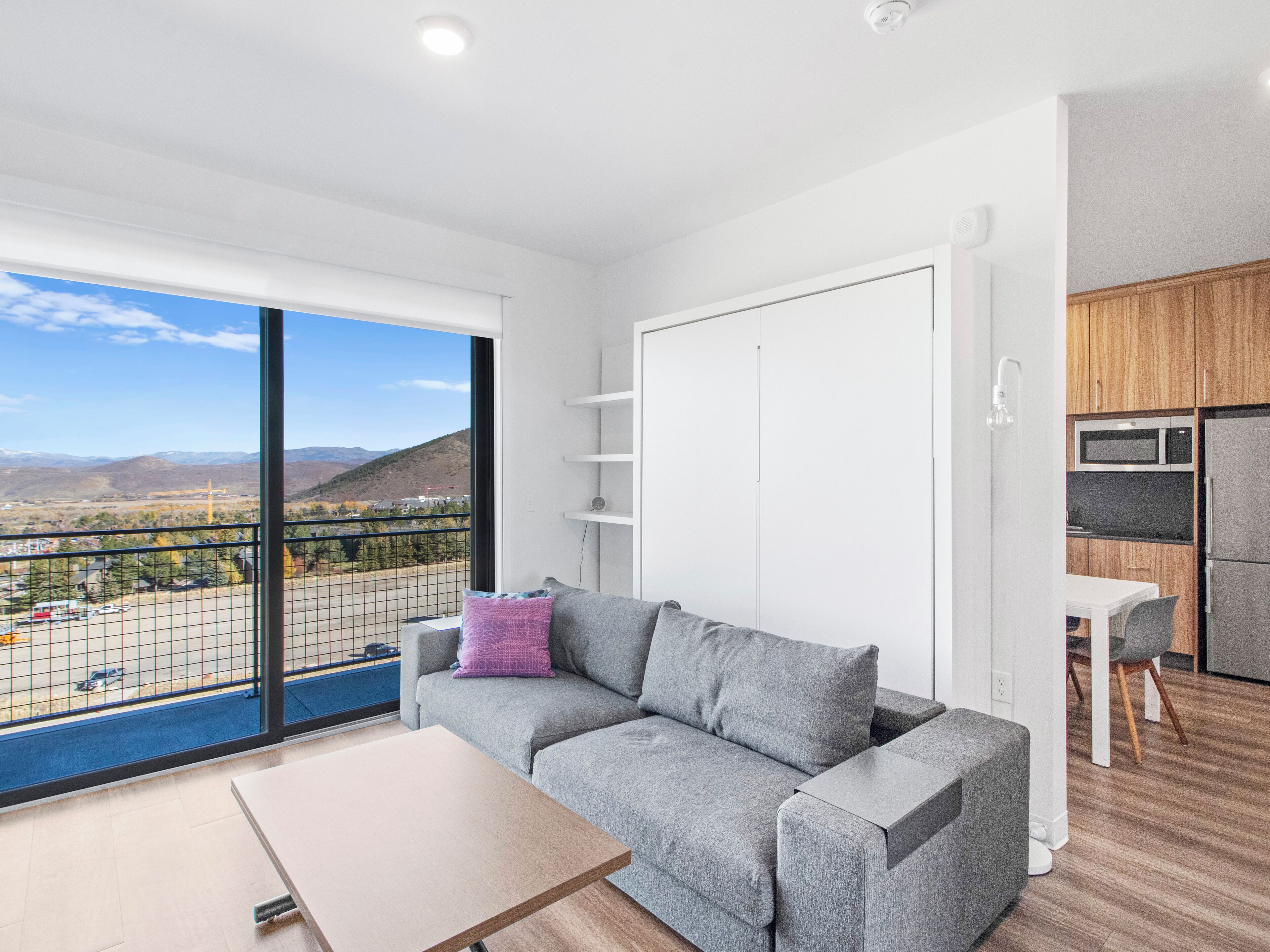A modern living room with a grey sofa, a small table, and a balcony with a view of mountains and a construction site.