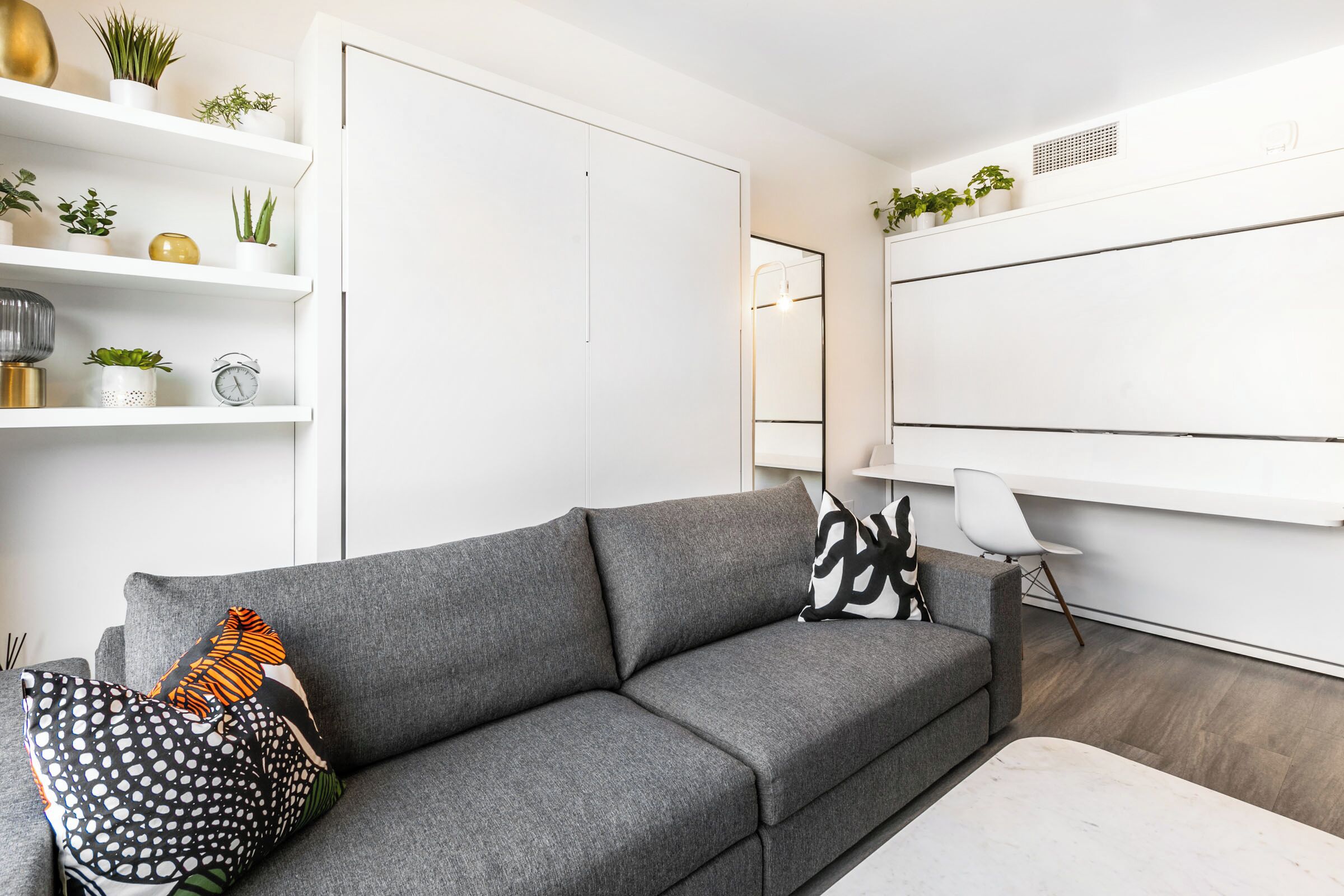 A modern living room with a grey sofa, a white coffee table, and a white chair. There are shelves with plants and decorative items.